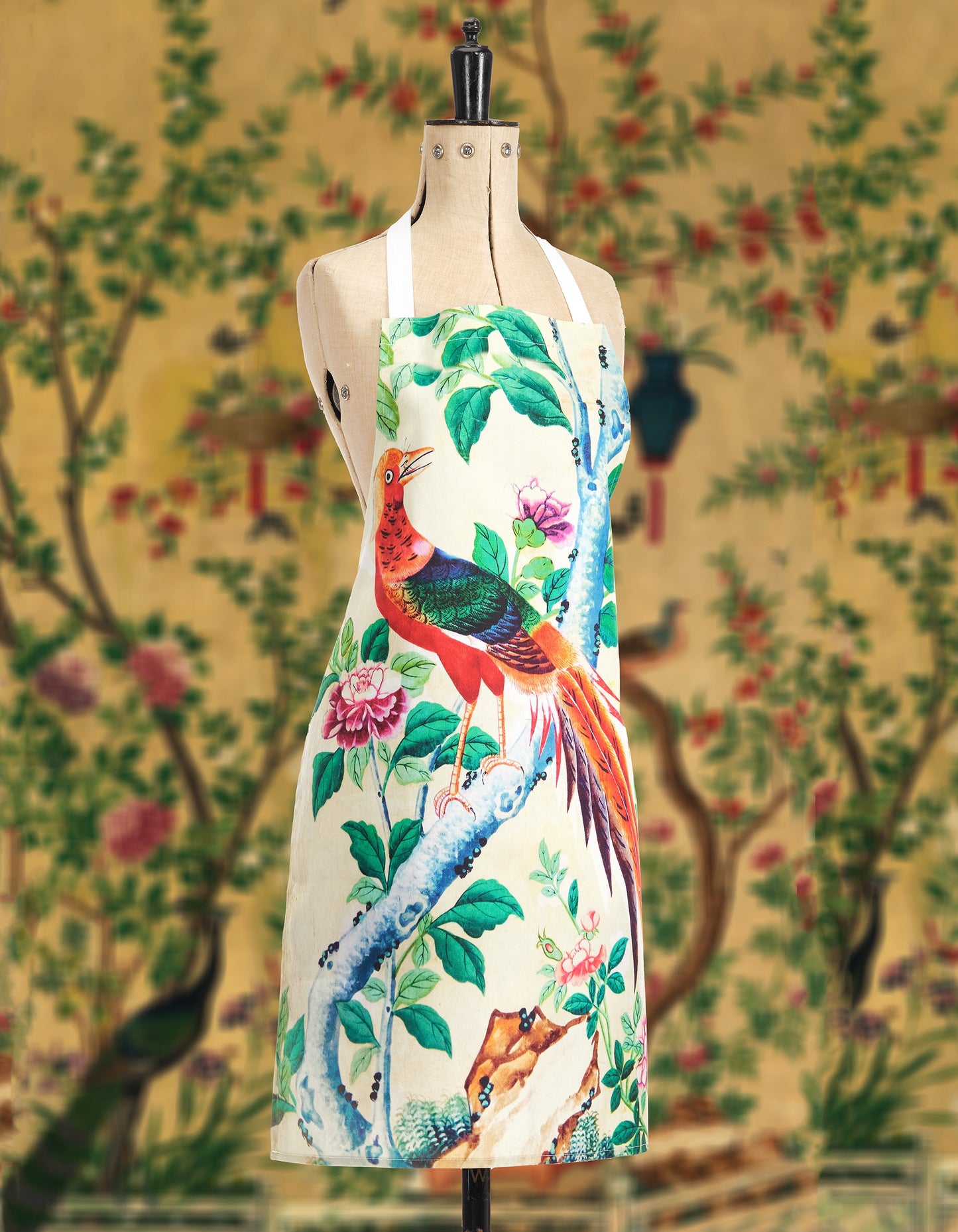 A mannequin wearing a pastel yellow apron decorated with birds, flowers and leaves in front of a matching yellow background.